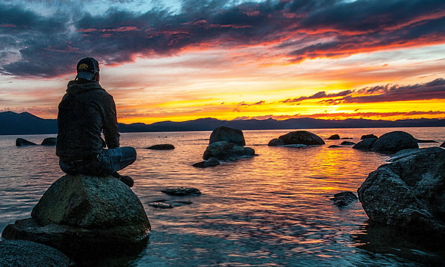 Ein Mann sitzt auf einem Stein und schaut sich den Sonnenuntergang am Wasser an.